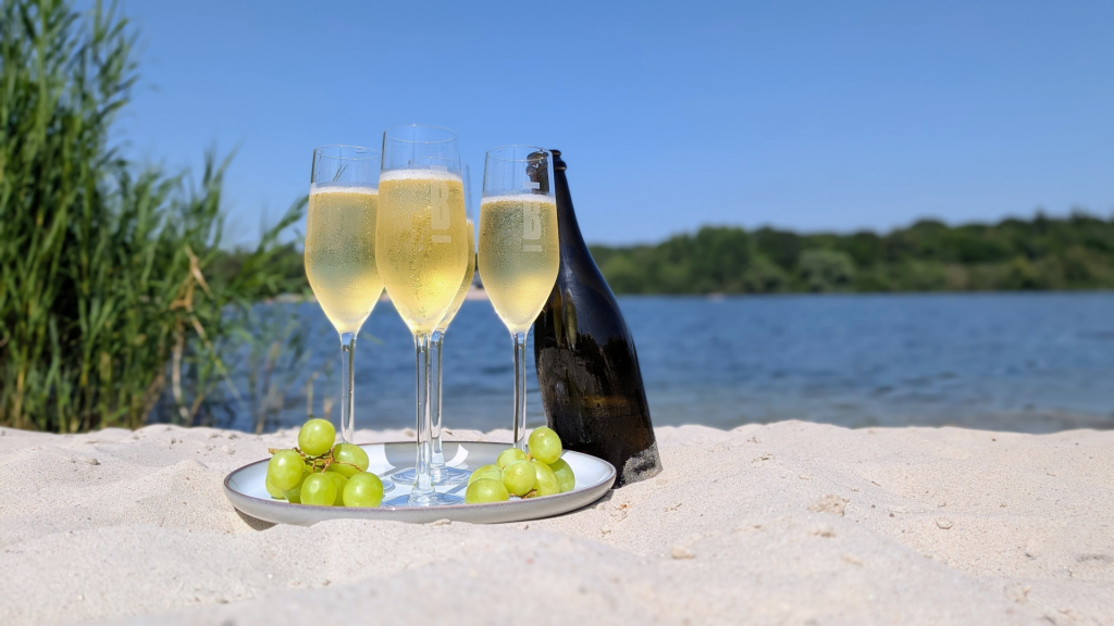 Auf dem feinen Sand eines Strands stehen Gläser mit eiskaltem Sekt, dazu knackige Trauben. Die Sonne strahlt. Kondenswasser bildet sich an den Gläsern.