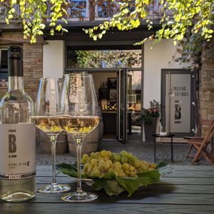 Auf einem Tisch sind zwei gefüllte Weingläser mit Buchert Wein und erntefrischen Trauben angerichtet. Der Hintergrund zeigt den malerischen Winzerhof von Weingut Buchert in Neustadt an der Weinstraße