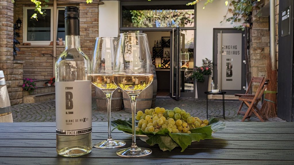 Auf einem Tisch sind zwei gefüllte Weingläser mit Buchert Wein und erntefrischen Trauben angerichtet. Der Hintergrund zeigt den malerischen Winzerhof von Weingut Buchert in Neustadt an der Weinstraße